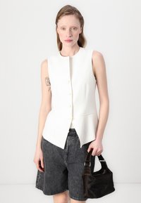 Hvit ermløs blazer med høy hals og knappelukking, kombinert med mørke denimshorts og en liten brun veske.