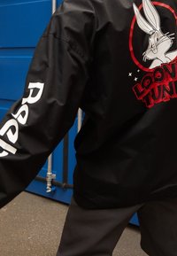 Person med svart jacka med Bugs Bunny och "Looney Tunes"-text på ryggen, vit text på ärmen, som står nära en blå vägg.