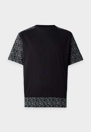 Ung mand stående iført en sort T-shirt og bukser med grå mønstrede sektioner og hvide sneakers mod en ensfarvet baggrund.