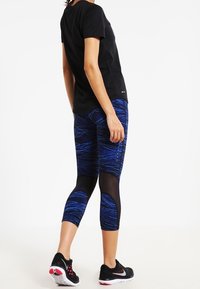 T-shirt nera a girocollo, leggings capri blu e neri con motivi e dettagli in rete, abbinati a scarpe sportive nere con dettagli rossi.