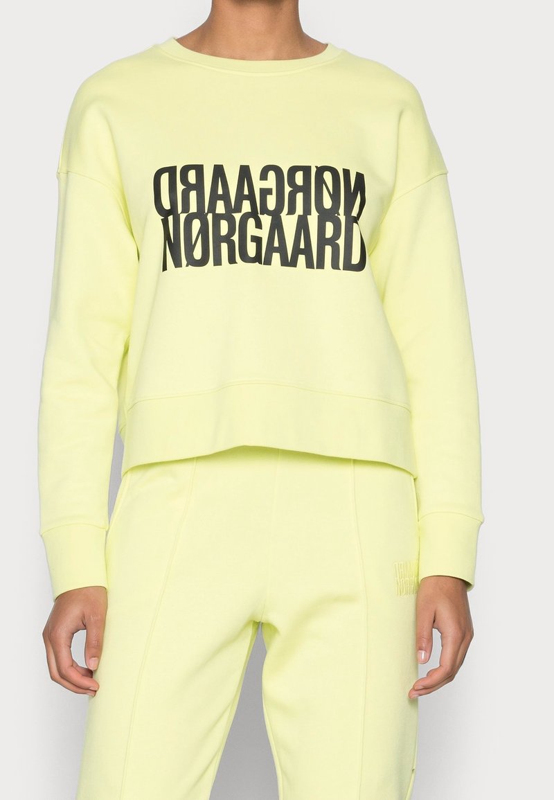 Personne portant un sweat-shirt et un pantalon jaunes pâles assortis ; le sweat-shirt présente un texte noir en gras « NØRGAARD » en miroir au-dessus du texte normal.