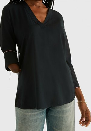 Mujer con blusa negra holgada de cuello en V con mangas tres cuartos y vaqueros de mezclilla azul claro, de pie frente a un fondo liso.