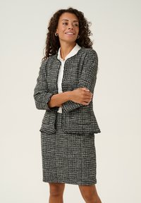 Fekete-fehér tweed öltöny, gallér nélküli zakóval, fodros szegéllyel és illeszkedő szoknyával, mindkettő textúrált, kockás mintával.