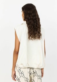 Top alb fără mâneci, cu o croială lejeră, având guler și tiv strâns. Împreunat cu pantaloni bej cu un model. Părul ondulat este vizibil.