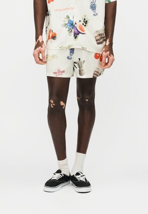 Άνδρας με λευκά σορτς και ταιριαστό πουκάμισο με πολύχρωμα σχέδια θερινών διακοπών, λευκές κάλτσες και μαύρα αθλητικά παπούτσια σε λευκό φόντο.