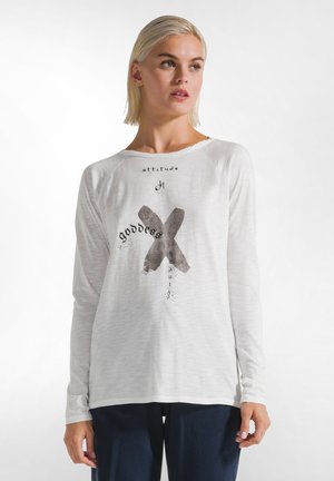 Langärmliges weißes Oberteil aus leichtem Stoff, versehen mit einem grafischen Druck mit Text und einem stilisierten schwarzen "X" auf der Vorderseite.