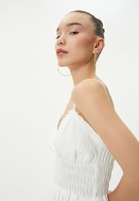 Abito bianco texturizzato con spalline sottili e corpino arricciato. Il modello indossa grandi orecchini a cerchio dorati. Lo sfondo è semplice e chiaro.