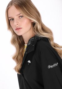 Jachetă de ploaie neagră cu guler lucios, logo umbrelă pe piept și text brodat pe mânecă. Textură transparentă și design cambrat.