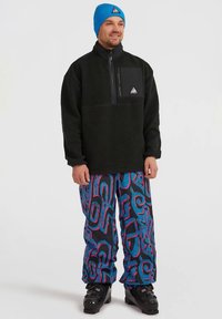 Mann in einer schwarzen Fleecejacke, blauer Strickmütze, bunten Hosen mit abstraktem Muster und schwarzen Skischuhen, der vor einem einfarbigen Hintergrund steht.