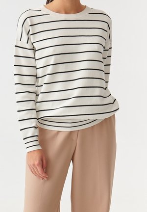 Pullover leggero a maglia in crema con strisce orizzontali nere. Caratterizzato da un collo rotondo e polsini a coste; abbinato a pantaloni larghi color cammello.