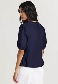 Blusa blu navy con maniche a palloncino, caratterizzata da un design arricciato laterale e chiusura con bottoni sul retro. Texture morbida e liscia con una vestibilità rilassata.