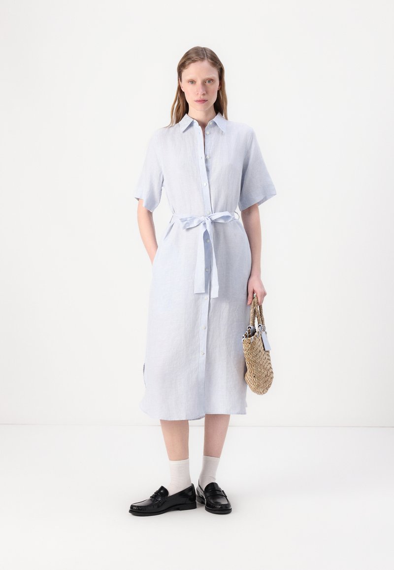 Hellblaues Leinenhemdkleid mit kurzen Ärmeln, Knopfleiste und gebundenem Taillengürtel. Getragen mit schwarzen Slippern und weißen Socken, während eine geflochtene Tasche gehalten wird.