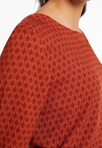 Gros plan sur une personne portant un vêtement rouge avec un motif géométrique orange en forme de losange et un col arrondi.