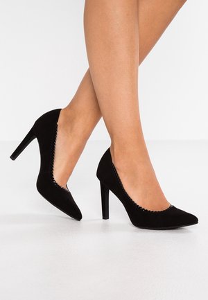 Modelo que lleva tacones altos de ante negro con punta puntiaguda y bordes festoneados, de pie sobre un suelo blanco contra un fondo blanco sencillo.