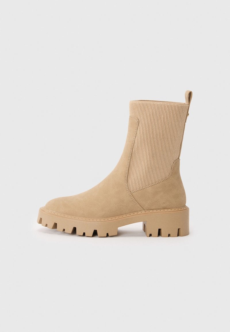 Botte beige mi-mollet avec semelle à crampons épaisse, panneau élastique côtelé, bout rond et languette à l'arrière, vue de profil sur un fond uni.