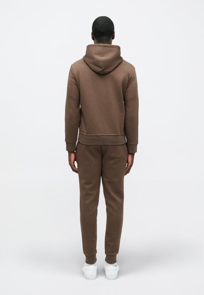 Smeđa dukserica i hlače od trenirke u mekom materijalu, s kapuljačom, rebrastim manžetama i opuštenim krojem. Bijele tenisice dodaju kontrast.