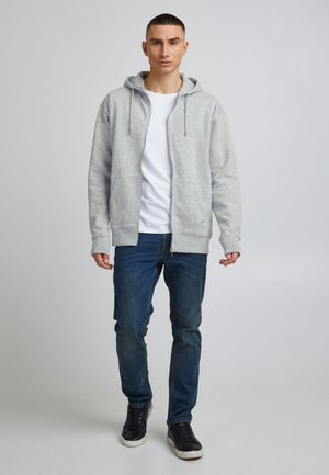 Grijze hoodie met ritssluiting en trekkoorden, gecombineerd met een wit T-shirt en blauwe jeans. Heeft twee voorkanten zakken en geribbelde manchetten.