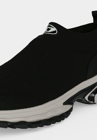 Μαύρο slip-on αθλητικό παπούτσι με υφασμάτινο επάνω μέρος με υφή, λευκή μεσαία σόλα και μαύρη ανθεκτική εξωτερική σόλα, που διαθέτει ασημένιο λογότυπο στο μπροστινό πάνω μέρος.