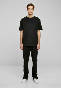 Černý bavlněný outfit skládající se z volného trička a ladících kalhot, obojí s jemnou texturou. Modelka stojí na jednobarevném pozadí.
