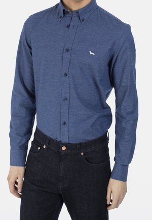 Mężczyzna w niebieskiej koszuli z długim rękawem zapiętej na guziki z małym białym logo psa na piersi oraz w ciemnych spodniach na białym tle.