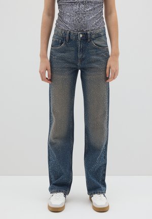 Donkerblauwe denim jeans met een ontspannen pasvorm, voorzien van lichte beschadigde accenten en verspreide witte stippen. Gecombineerd met witte sneakers.