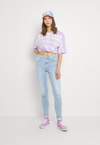 Joven mujer vistiendo una camiseta corta de tirantes anudada de tie-dye lavanda, jeans skinny de color azul claro, zapatillas rosas y una gorra de béisbol colorida, contra un fondo blanco.