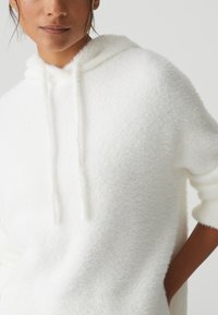 Weiße, flauschige Kapuzenjacke aus strukturiertem Stoff. Verfügt über eine Kordelzugkapuze und einen lässigen Schnitt, mit 3/4-langen Ärmeln und seitlichen Taschen.