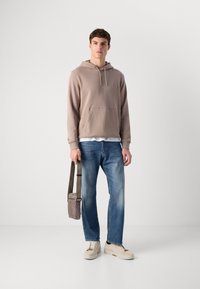 Jonge man die staat in een casual beige hoodie, blauwe jeans en lichte sneakers, terwijl hij een kleine taupe schoudertas bij de hengsel vasthoudt.