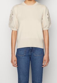Béžový pletený svetr s krátkými nabíranými rukávy, který má detaily v podobě průstřihů. V kombinaci s modrými džíny, zdůrazňující ležérní vzhled.