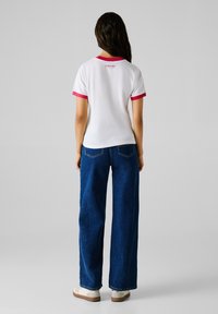 Tricou alb cu mânecă scurtă și detalii roz, purtat cu blugi largi de culoare albastru închis și adidași albi. Logo cu text pe spate.
