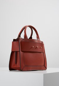 Sac à main en cuir rouge avec un design structuré, comprenant une poche avant, trois clous décoratifs en or et deux poignées rondes.