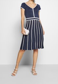 Marinblå klänning med V-ringning, korta ärmar och vita vertikala ränder, tillverkad av ett texturerat tyg; matchad med en pärlklädd clutch och remsandaler.