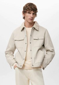 Beige Jacke mit Sherpa-Kragen, Druckknöpfen und zwei Brusttaschen. Der Stoff wirkt robust und das Design sauber. Über einem leichten Pullover getragen.