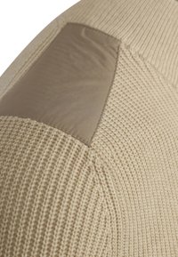 Gehaakte beige trui met een gestructureerde afwerking, voorzien van een gladde tan vlek op de schouder, die de contrasterende materialen en het ontwerp benadrukt.