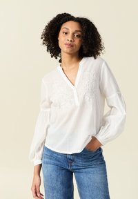 Blusa bianca a maniche lunghe con scollatura a V e dettagli floreali ricamati sul petto. Presenta maniche arricciate e un tessuto leggero.