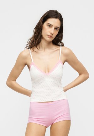 Νεαρή γυναίκα με μακριά κυματιστά μαλλιά, φοράει λευκό τοπ με ροζ καρδιές και ροζ σορτς, στέκεται με τα χέρια στη μέση.