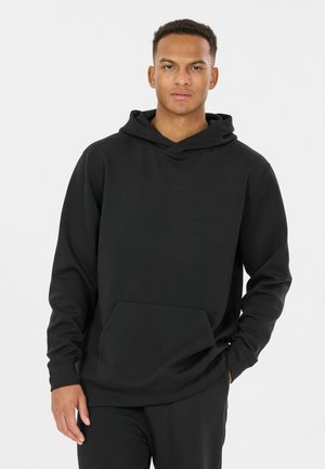 Young man standing wearing a black hooded sweatshirt with a front pocket and black pants against a plain light background.