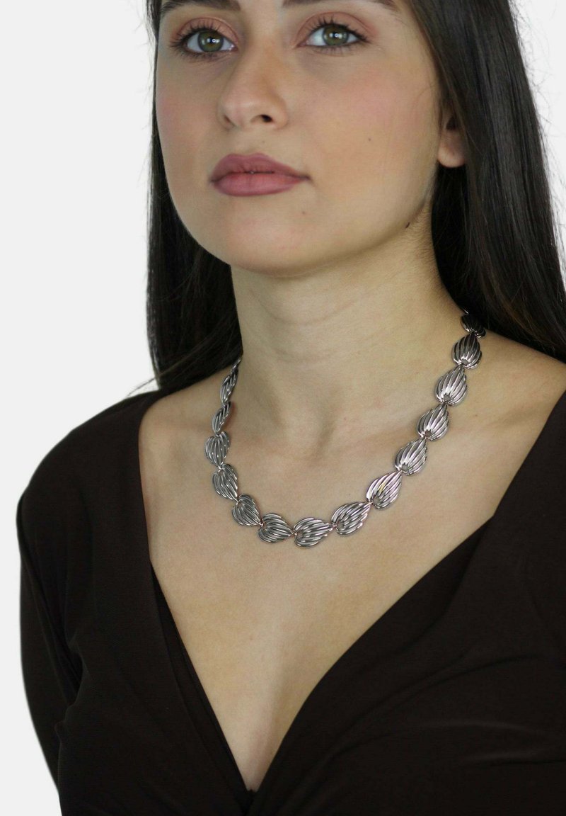 Collar de metal plateado con eslabones en forma de hojas entrelazadas y ondulantes. La cadena tiene un acabado brillante y se lleva con una blusa de escote en V profundo.