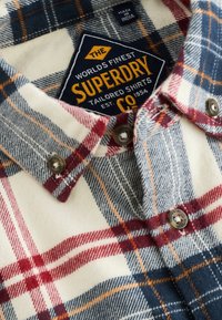 Flannelskjorte med ternet mønster i rød, blå og creme. Den har metalknapper og et mærke med "Superdry" branding.