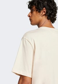 T-shirt in cotone beige con vestibilità relaxed, maniche corte e scollatura rotonda, mostra la vista posteriore e una texture liscia, design minimalista.