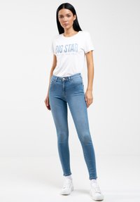 Jeans ajustados de denim azul con cintura alta y diseño de cinco bolsillos, combinados con una camiseta blanca que tiene texto en azul claro. Zapatillas blancas.