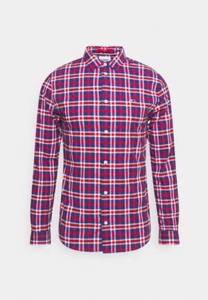 Chemise à manches longues boutonnée avec un motif à carreaux rouge, bleu et blanc et un petit logo sur la poitrine gauche.