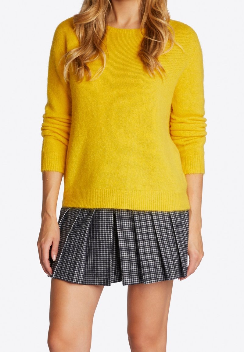 Femme portant un pull jaune vif à manches longues et une mini-jupe plissée à carreaux noir et blanc, debout, les bras détendus.