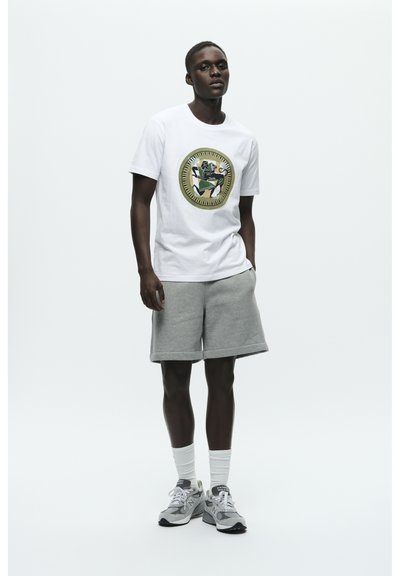 Biały bawełniany t-shirt z okrągłym nadrukiem z kolorowym wzorem. Zestawiony z szarymi szortami z polaru, białymi skarpetkami i szarymi trampkami.