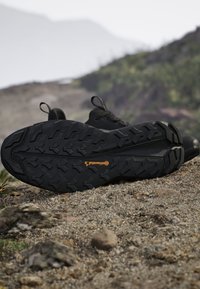 Scarpa da trekking nera posizionata con la suola verso l'alto su terreno roccioso, mostrando un battistrada robusto con marchio Continental, all'aperto con colline sfocate sullo sfondo.