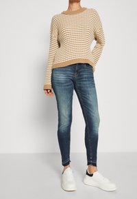 Suéter de punto a rayas beige y blanco, combinado con jeans ajustados azules y zapatillas blancas con toques negros. Presenta un corte relajado y textura acanalada.