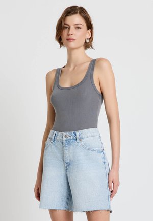 Donna che indossa una canotta grigia a costine e pantaloncini di jeans azzurri a vita alta con orli sfrangiati, in piedi davanti a uno sfondo semplice.