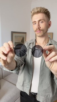Okrogle črne sončne očala s temnimi lečami in tankim kovinskim okvirjem, ki so jih držali v dveh rokah proti svetli podlagi in mehko teksturirani kavču.