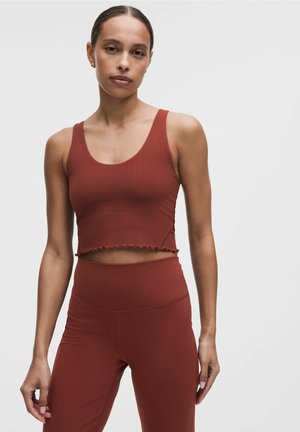 Kvinde med håret sat op i en knold iført en rustfarvet ribstrikket kort tanktop og matchende leggings med høj talje, stående mod en ensfarvet hvid baggrund.