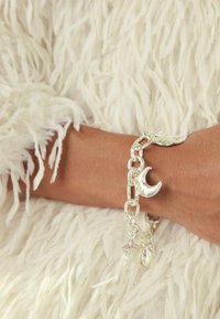 Sølv kædearmbånd med måne-, stjerne- og bladcharms, sat mod en tekstureret hvid pelsbaggrund. Glat overflade, reflekterende finish.